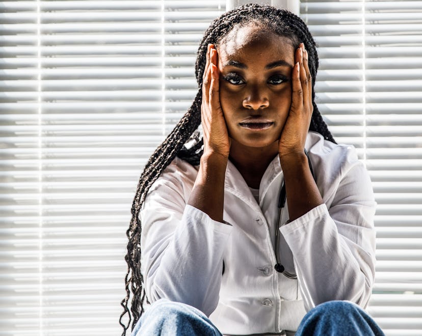 Portrait of overworked female doctor sitting on the floor, by the window, head in hands, looking at ...