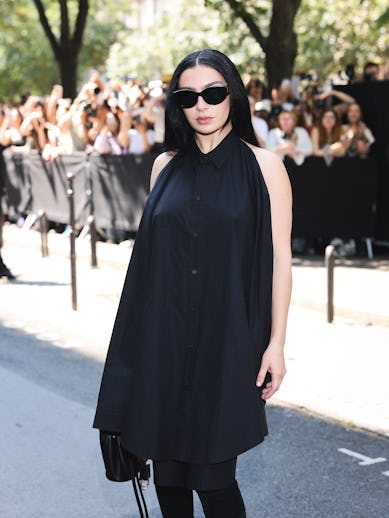 A woman in sunglasses and a black dress poses at an event, with a crowd and security barriers in the...