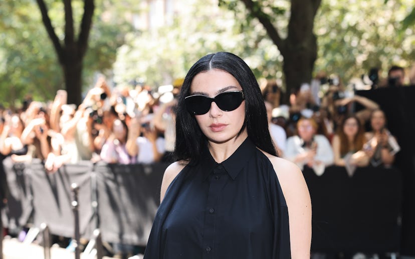 A woman in sunglasses and a black dress poses at an event, with a crowd and security barriers in the...