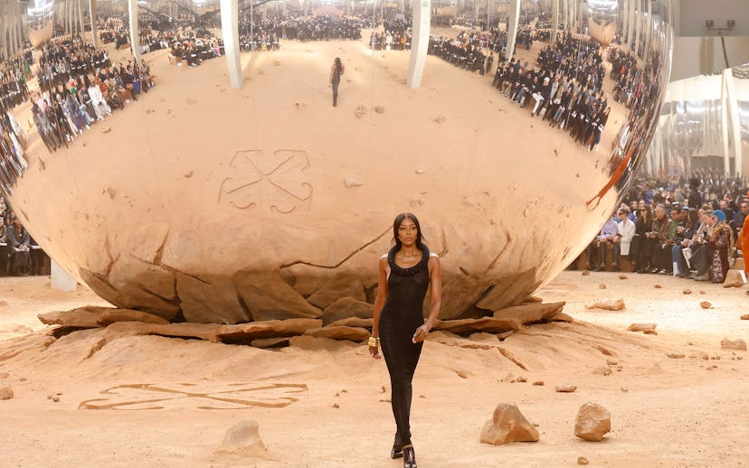 A model walks on a runway in front of a large mirrored sphere surrounded by a crowd, set on a sandy ...