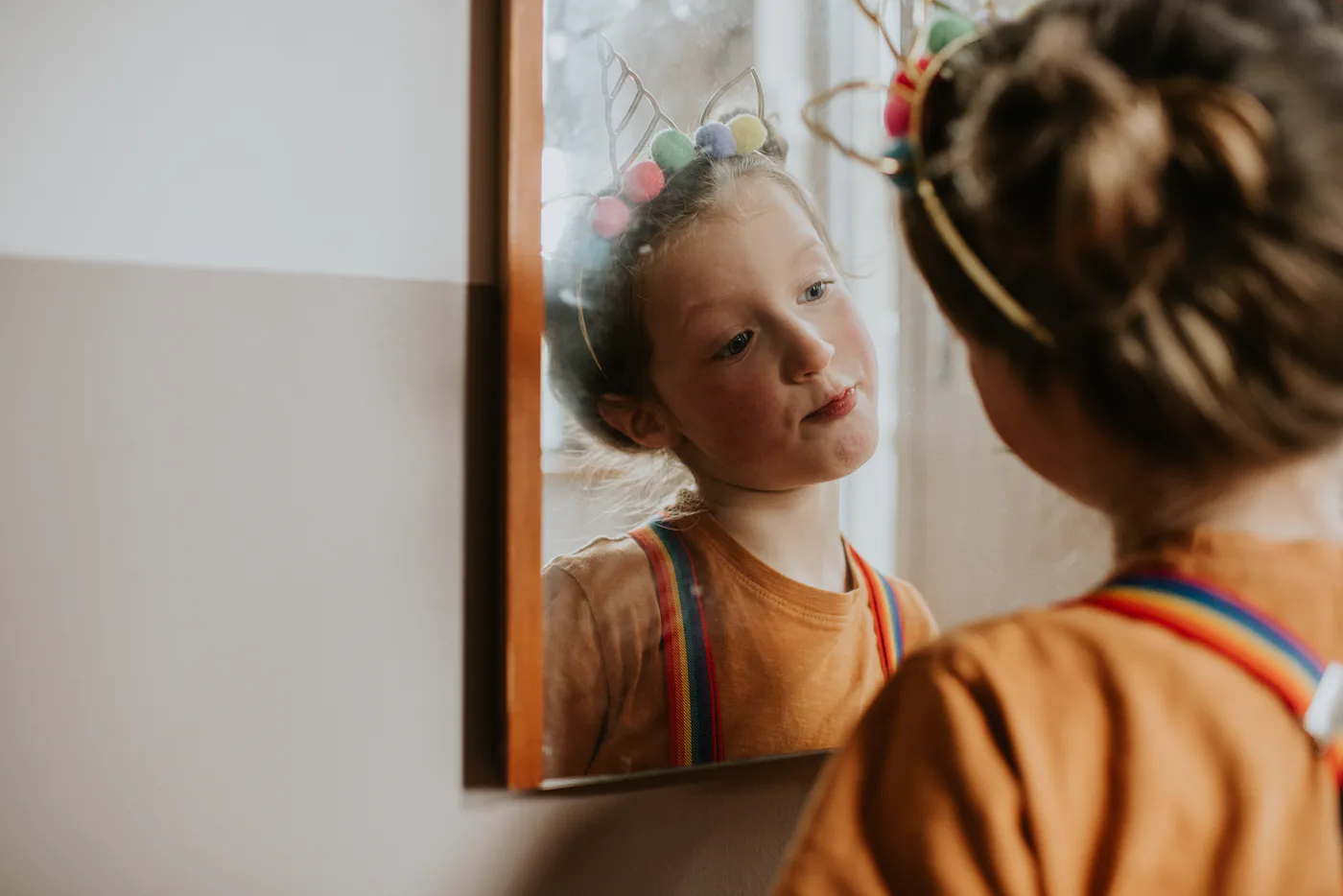 A young girl wearing a quirky unicorn headband and rainbow braces looks at her reflection in a mirror. She tilts her head in contemplation, and seems rather confident and self assured in her appearance and fashion sense.