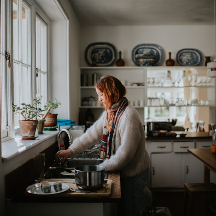 A young woman stands by a sink and washes dishes under a faucet.
