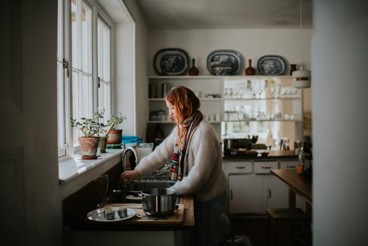 A young woman stands by a sink and washes dishes under a faucet.