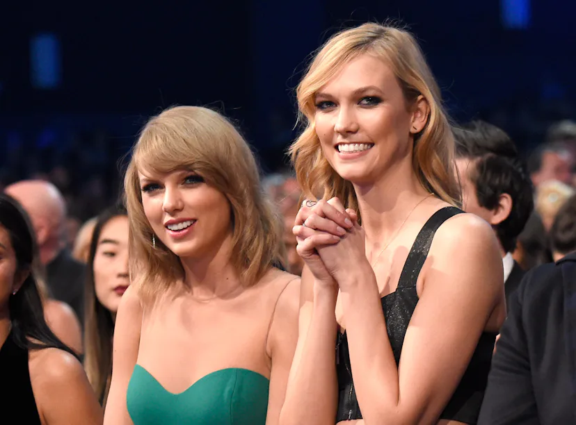 Two women in elegant dresses smiling at a formal event, one in a green strapless dress and the other...
