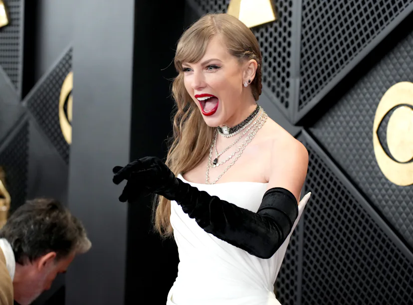 A woman in a white gown and black gloves laughing excitedly at an event. A man is seen bending down ...