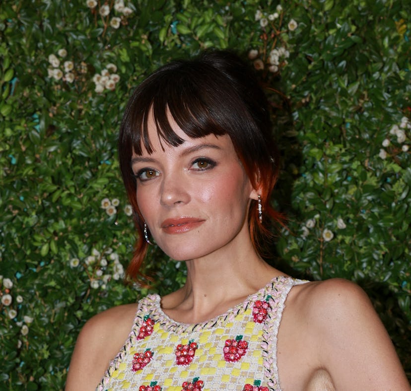 Woman with shoulder-length brown hair, wearing a floral patterned dress, stands in front of a leafy ...