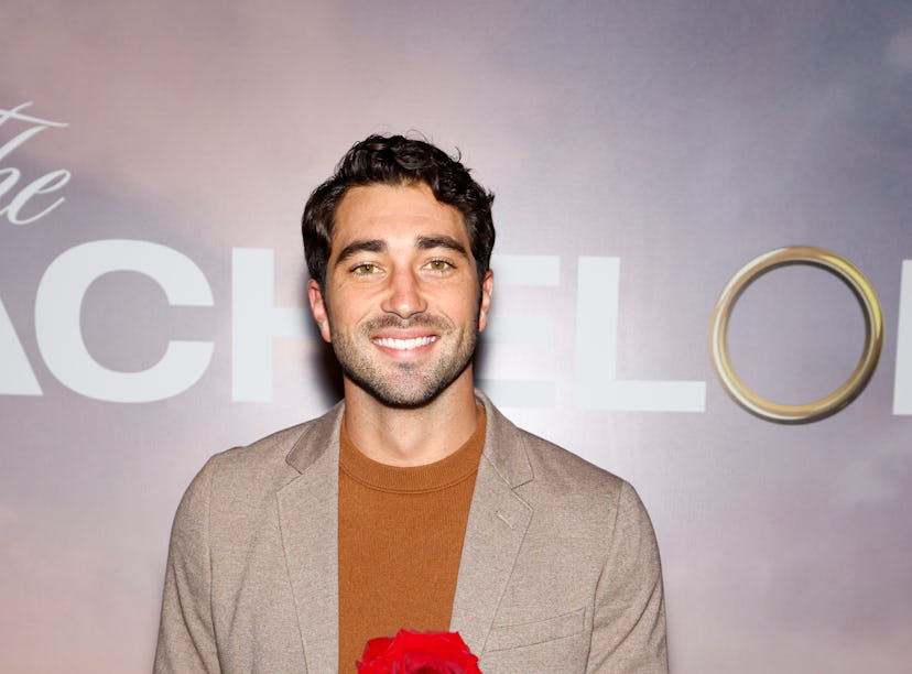 Man in a blazer and sweater smiling and holding a red rose in front of a "The Bachelor" themed backd...