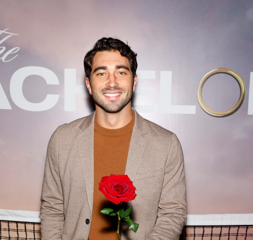 Man in a blazer and sweater smiling and holding a red rose in front of a "The Bachelor" themed backd...