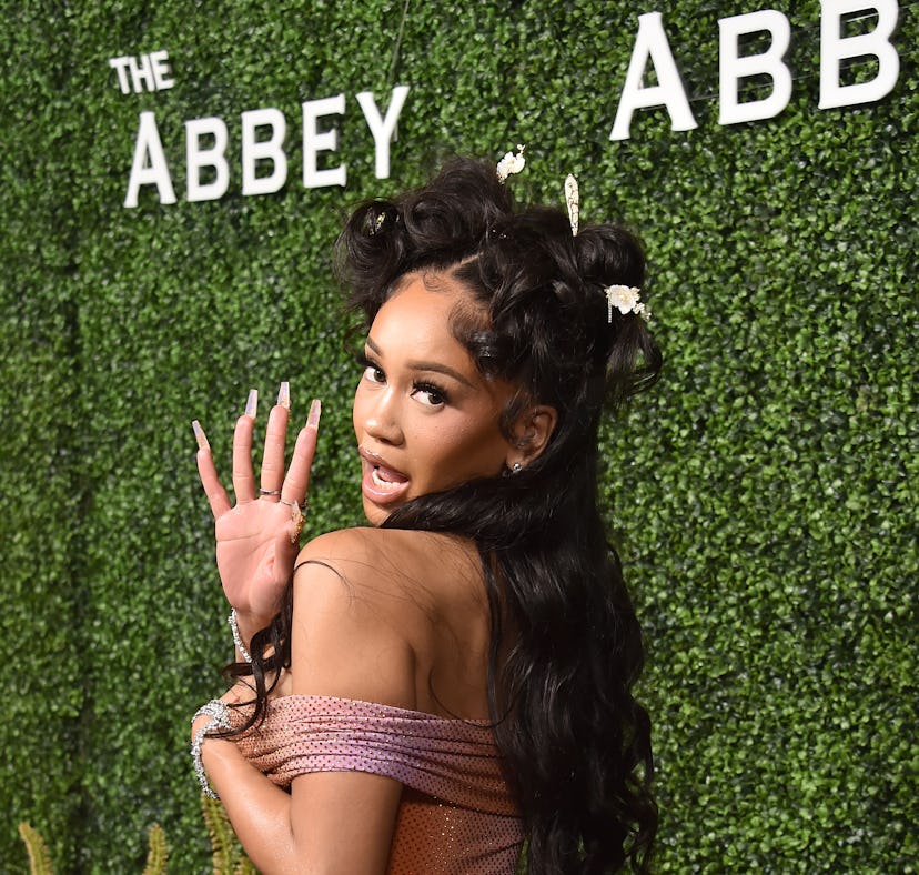 Woman in a sparkling peach dress posing playfully at an event, standing in front of a greenery wall ...