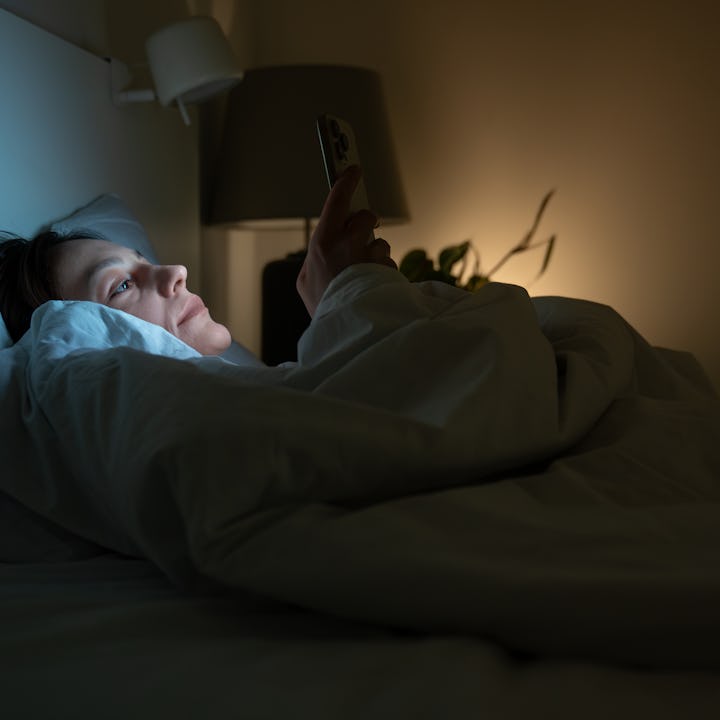 Exhausted woman with insomnia lying in bed in dark bedroom, looking at screen of smartphone. Female ...