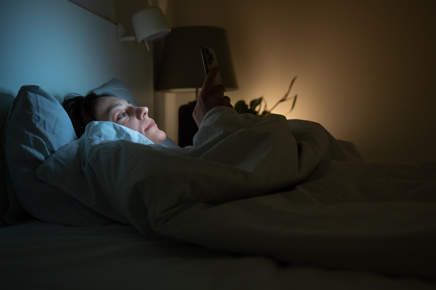 Exhausted woman with insomnia lying in bed in dark bedroom, looking at screen of smartphone. Female with sleeping disorder, anxiety. Mobile phone addiction influencing melatonin production at night