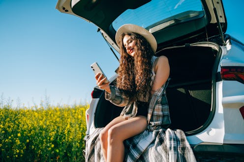 A happy brunette woman in a hat and shirt stops in a rapeseed field, using a smartphone while taking...