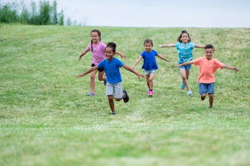 A small group of young children are seen running down a grassy hill with their arms out like airplan...