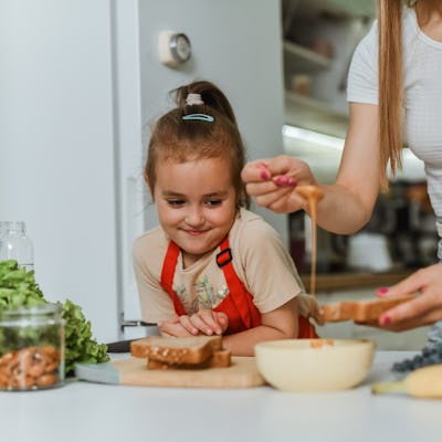 Female Child Looking Greedily At Mother Making Her Bread With Peanut Butter And Banana Slices