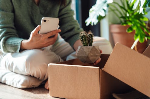 Young woman holding smartphone and taking out a small house plant from delivery box she purchased on...