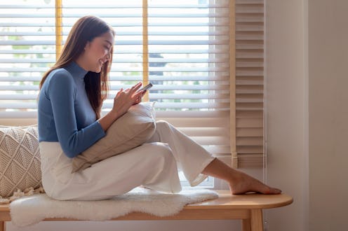 Smiling Asian cheerful adult female enjoy using smartphone surf internet social media relax casual d...
