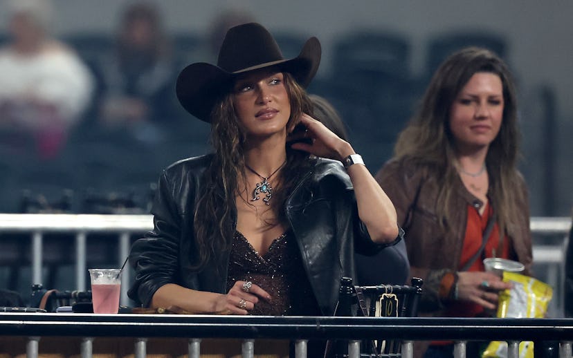 A woman in a black cowboy hat and leather jacket leans on a railing at a public event, looking thoug...
