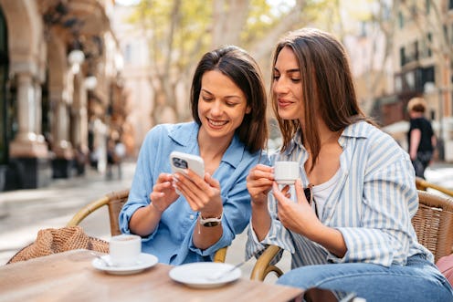 Two female friends using a phone while drinking coffee in a sidewalk café in Palma de Mallorca in Sp...