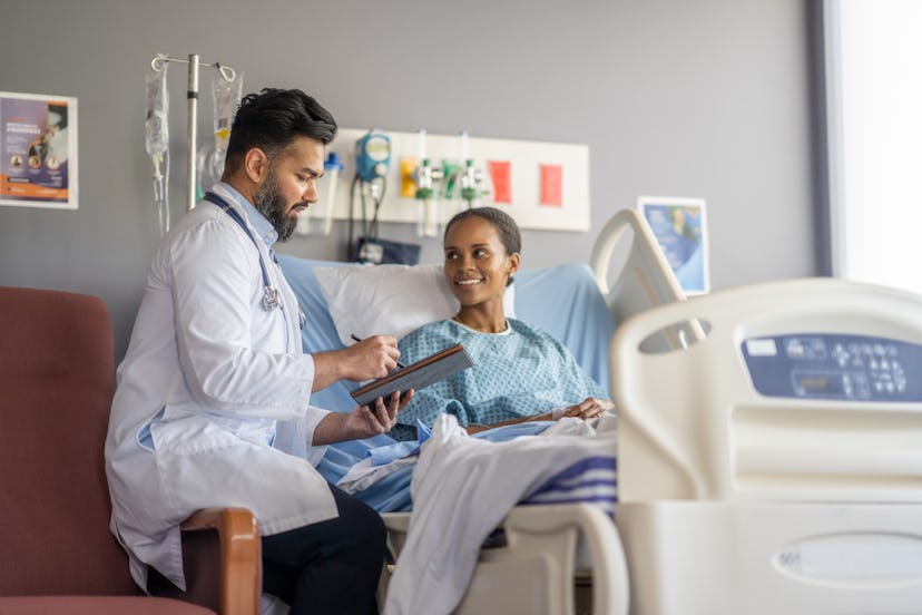 A woman sits up in a hospital bed wearing a gown as she talks with her male doctor about her surgery…