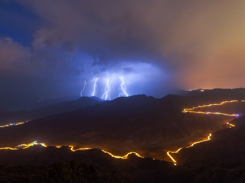 Lightning strikes illuminate a mountainous landscape at night, with winding roads lit by golden ligh...