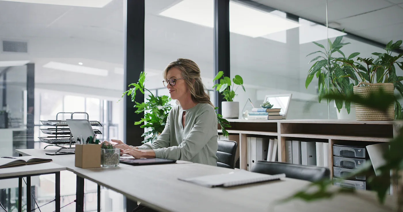 Laptop, typing and mature business woman at desk in glass office for administration or company management. Computer, research and report with confident mature manager in professional workplace