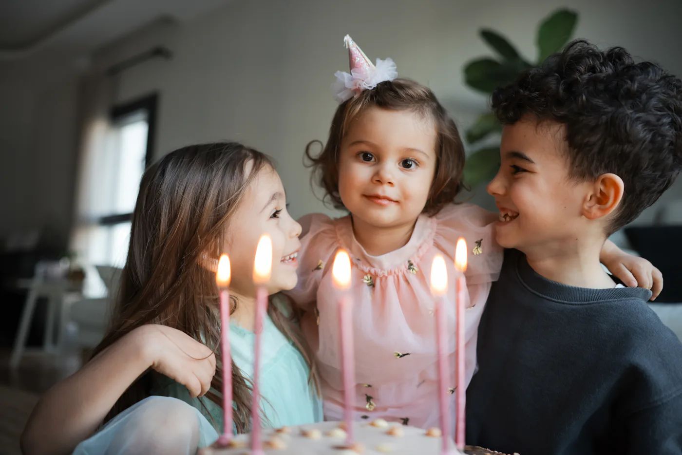 Child birthday party. Adorable little boy and girl with birthday cake decorated flowers and burn candle, makes wish. Kid in festive dress at room with balloons, bouquet of flowers at vase