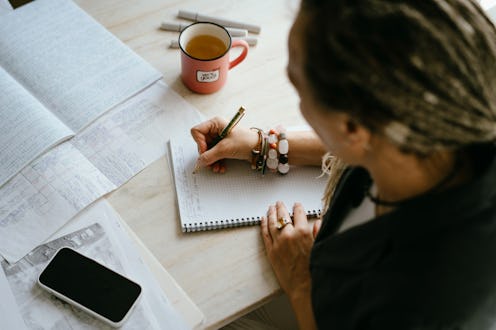 Portrait of a woman writing by hand with a pen in a large notebook, red cup of tea on the table, doc...