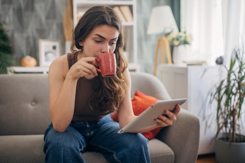 A woman relaxes with a cup of coffee while looking at a tablet, indicating a break or leisure activi...