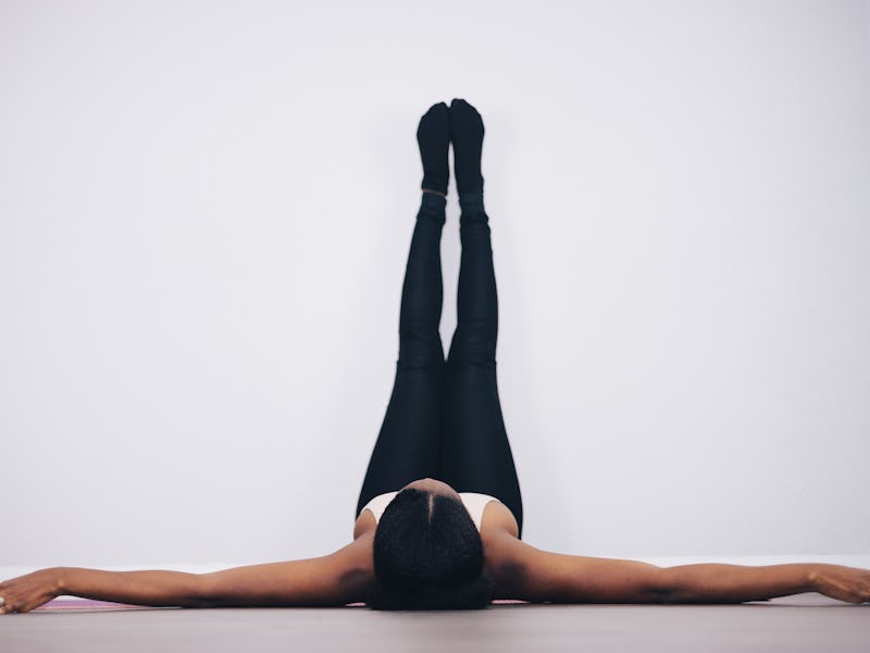 A person lying on a yoga mat with their arms outstretched and legs elevated against a white wall, we...