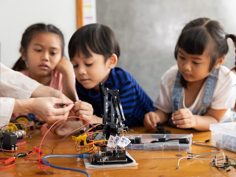 A group of young children intently focuses on a hands-on robotics activity, guided by their teacher,...