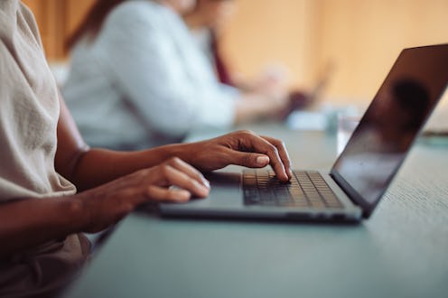 Close-up image of a person's hands typing on a laptop in an office environment with colleagues in th...