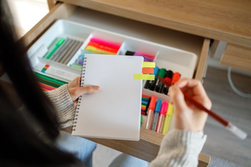 A person selects a marker from a drawer full of colorful stationery, holding a notepad with sticky n...