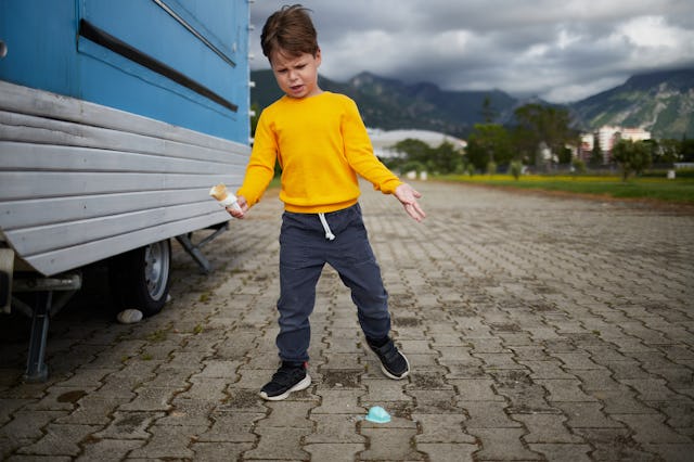 Young boy in yellow shirt looks upset after dropping his ice cream cone on the pavement near a blue ...