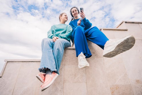 Two young female friends sitting with their phones on a staircase against the sky, view from below. ...