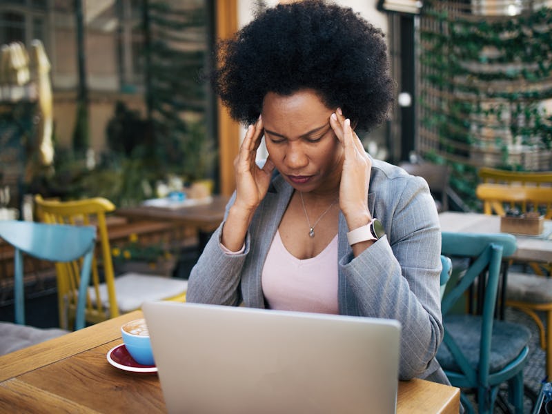 Black woman is sitting in a cafe and has a bad headache while working on a laptop