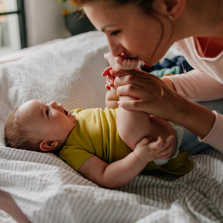 Portrait of a little smiling baby boy and his mom, kissing his tiny feet right after waking up in hi...
