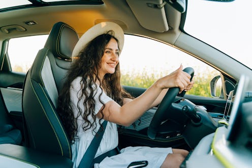 A smiling young brunette woman drives a car through a sunny rapeseed field in summer season