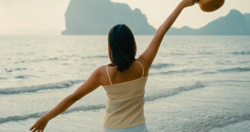 Young Asian woman walking barefoot along on sandy beach during sunset, enjoying tranquil waves and w...