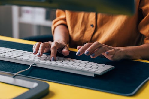 Close-up of a woman's hands typing on a white computer keyboard with a yellow desk, capturing a mome...