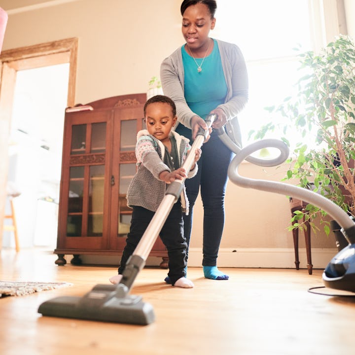 Cute little African girl helping her mom vacuum up their living room at home while doing household c...