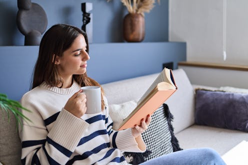 Woman is enjoying a relaxing afternoon at home, sitting on her sofa while reading a book and drinkin...
