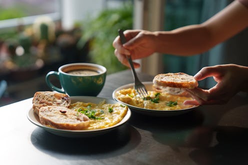 A cropped image of a woman's hand holding a piece of toasted bread with scrambled eggs on top, as sh...