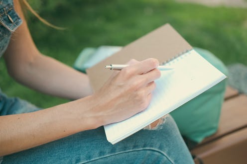 Close-up shot of cropped young woman in ripped knee denim overalls writing in note pad while sitting...