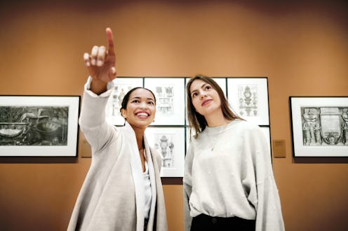 Two women smile and engage with art as one points out a detail in a gallery. The image captures a mo...