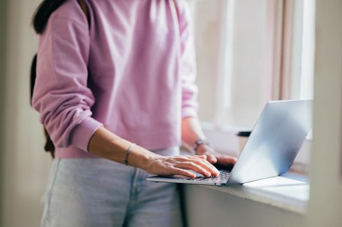 A woman in casual attire types on a laptop by a window, bathed in natural light, emphasizing modern ...