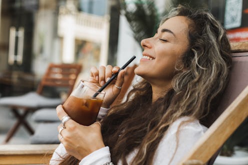 Cheerful girl drinking iced coffee outdoors. Positive emotions and laugh. Youth culture and cafe lif...