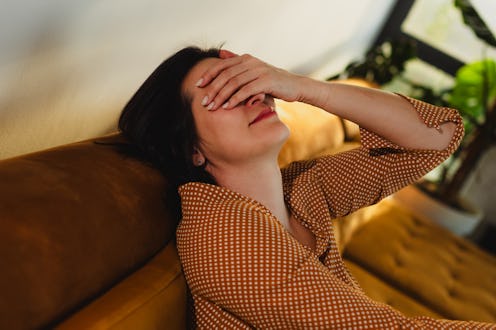 A woman in a patterned shirt sits on a couch with her hand covering her forehead, expressing stress ...