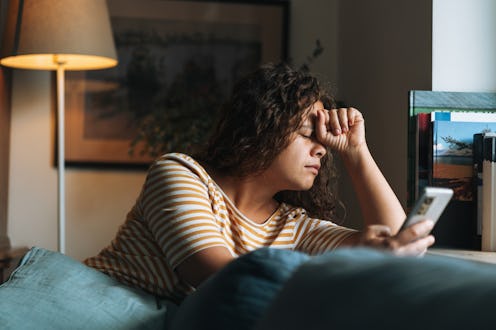 Depressed woman with curly hair covering face with hands using mobile phone sitting on sofa in room ...