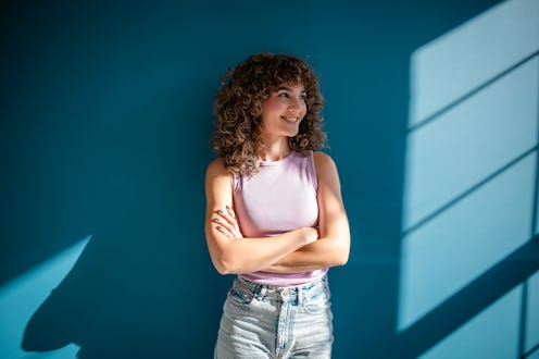 A cheerful young woman with curly hair and casual outfit poses confidently against a teal wall, illu...