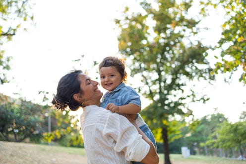 A smiling mother lifts her happy child in a sunny park, surrounded by lush green trees. The moment c...
